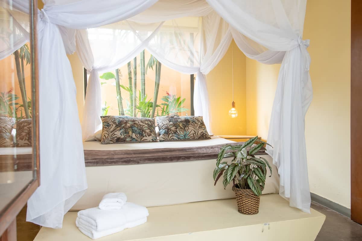 Ein Zimmer mit einem Bett, einem Vorhangfenster, einer Topfpflanze und einer Wandleuchte.