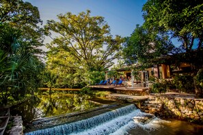 A serene outdoor setting with a waterfall, a stone bridge, and a building with blue chairs. - Hacienda Boutique  (Restrepo)