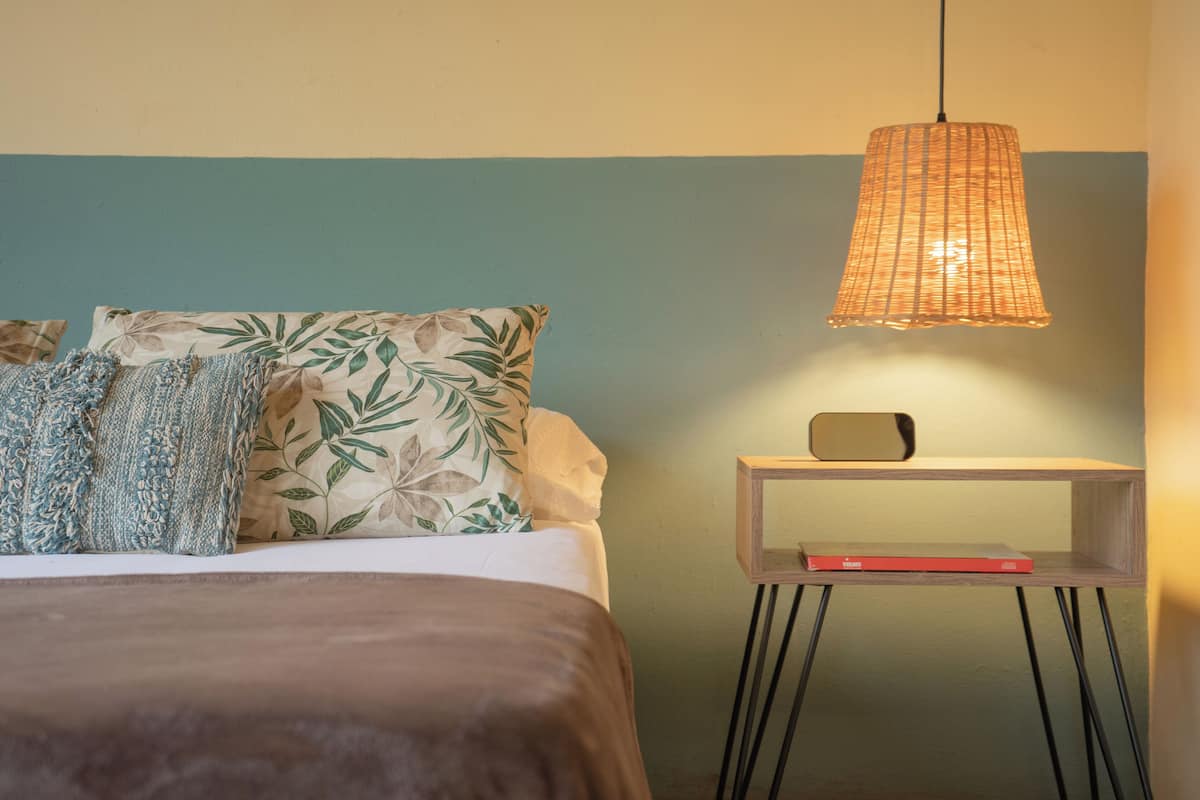 Ein ordentlich bezogenes Bett mit gemusterten Kissen, ein Nachttisch mit Lampe und eine kleine Ablage mit einem Buch.