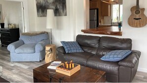 A living room with a brown leather sofa, a blue striped armchair, a wooden coffee table, and a guitar on the wall.