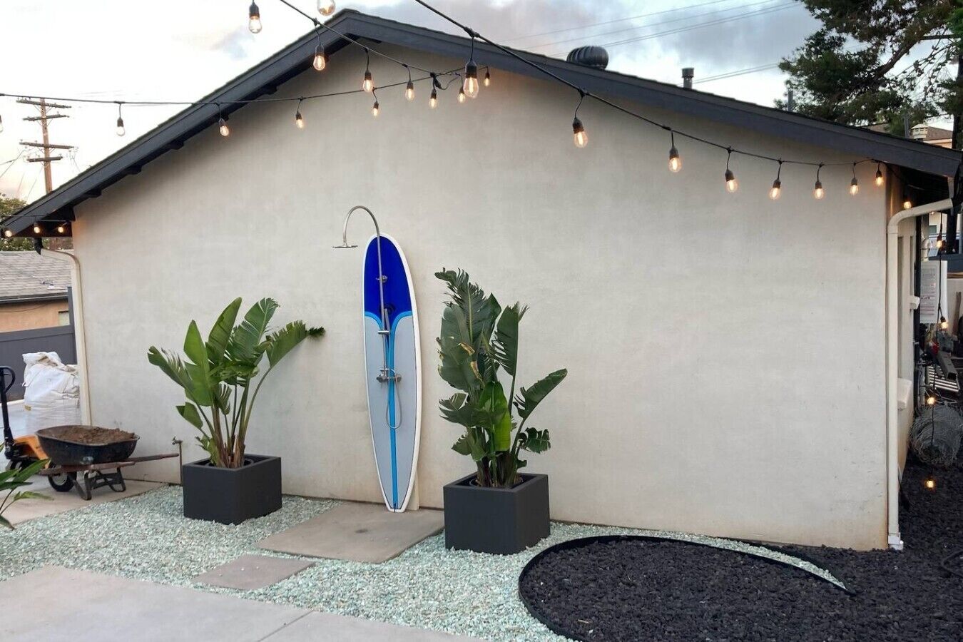A building with a surfboard mounted on the wall, potted plants, and string lights.
