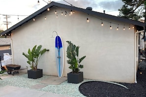 A building with a surfboard mounted on the wall, potted plants, and string lights.