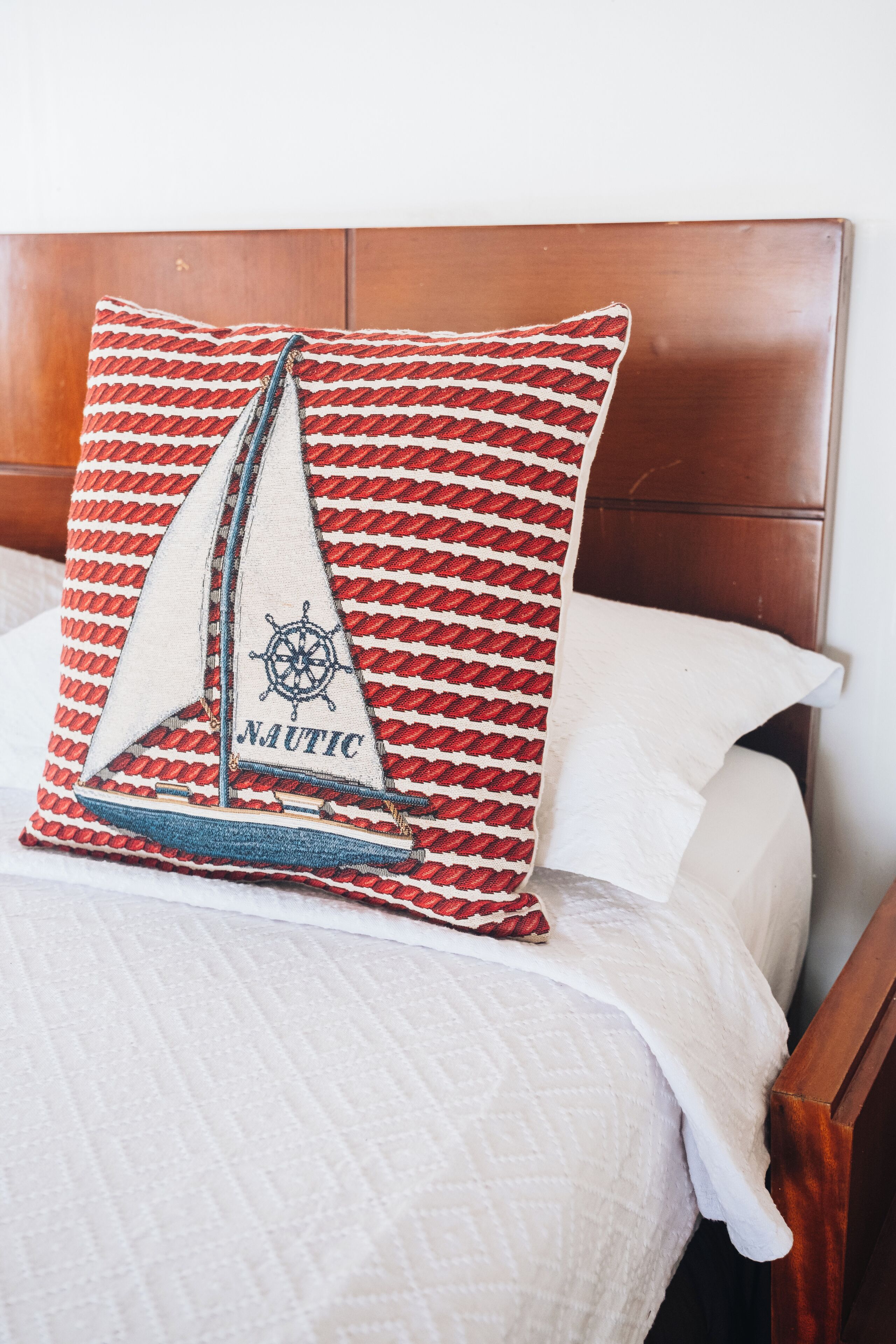 A bed with a wooden headboard, a quilted white bedspread, and a red and white patterned pillow with a sailboat design and the word "NAUTIC".