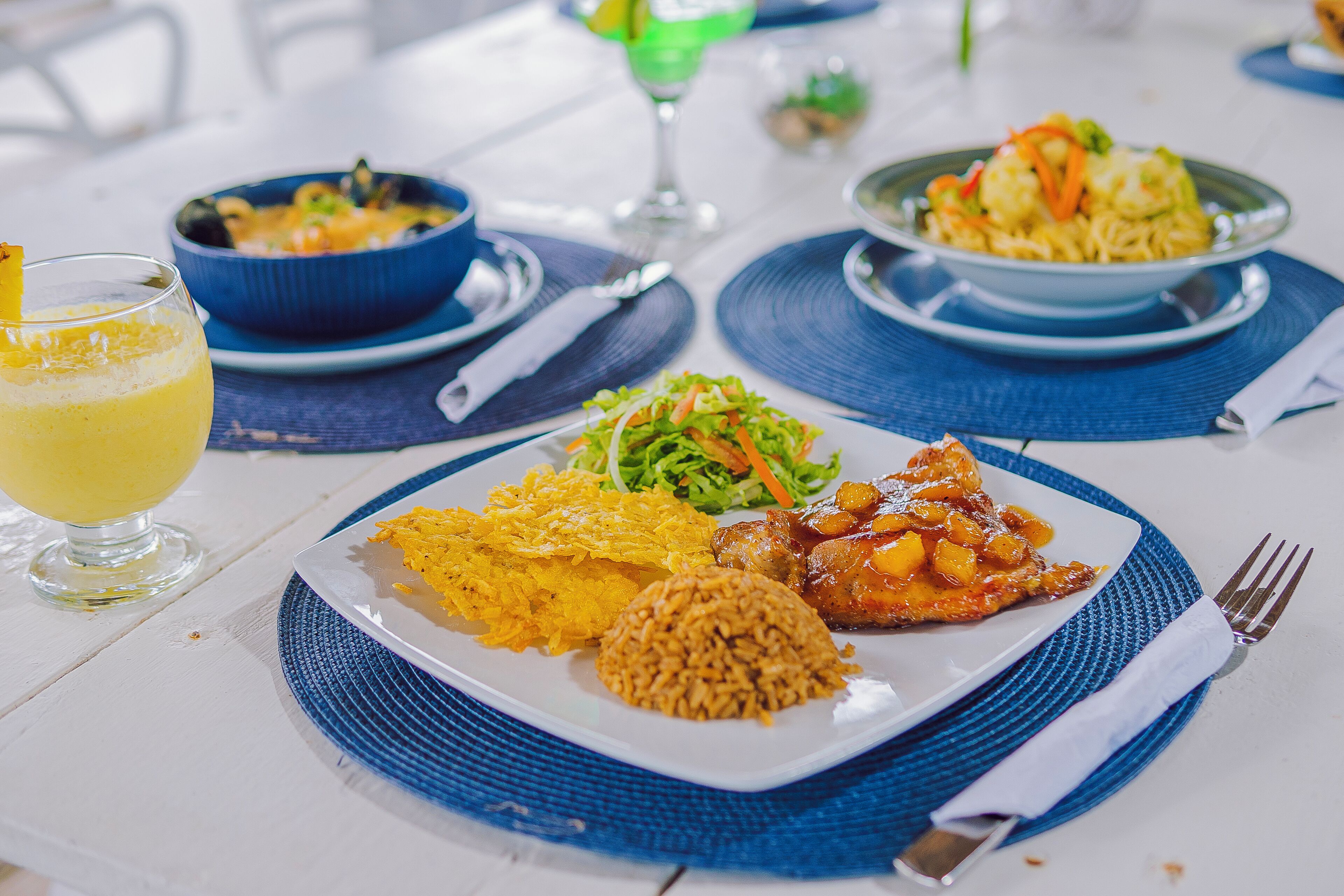 Una mesa de comedor con varios platos, incluyendo arroz, una ensalada y un guiso de carne, junto con un vaso de una bebida amarilla.
