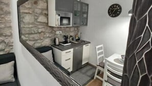 Una cocina compacta con gabinetes blancos, un microondas y un lavamanos. Hay una zona de comedor con una mesa y sillas blancas. Se ve un reloj en la pared.