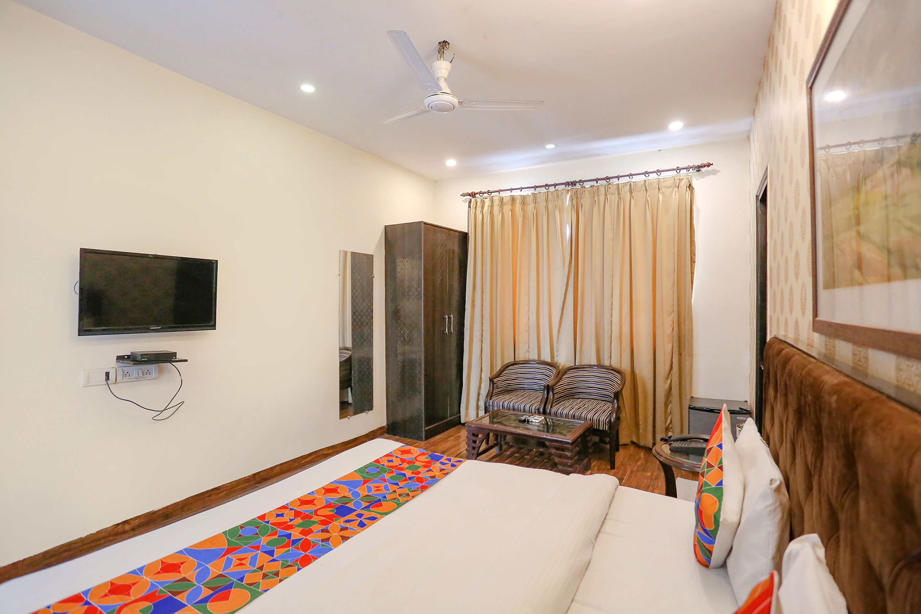 A hotel room with a bed, a television mounted on the wall, a ceiling fan, and a window with curtains.