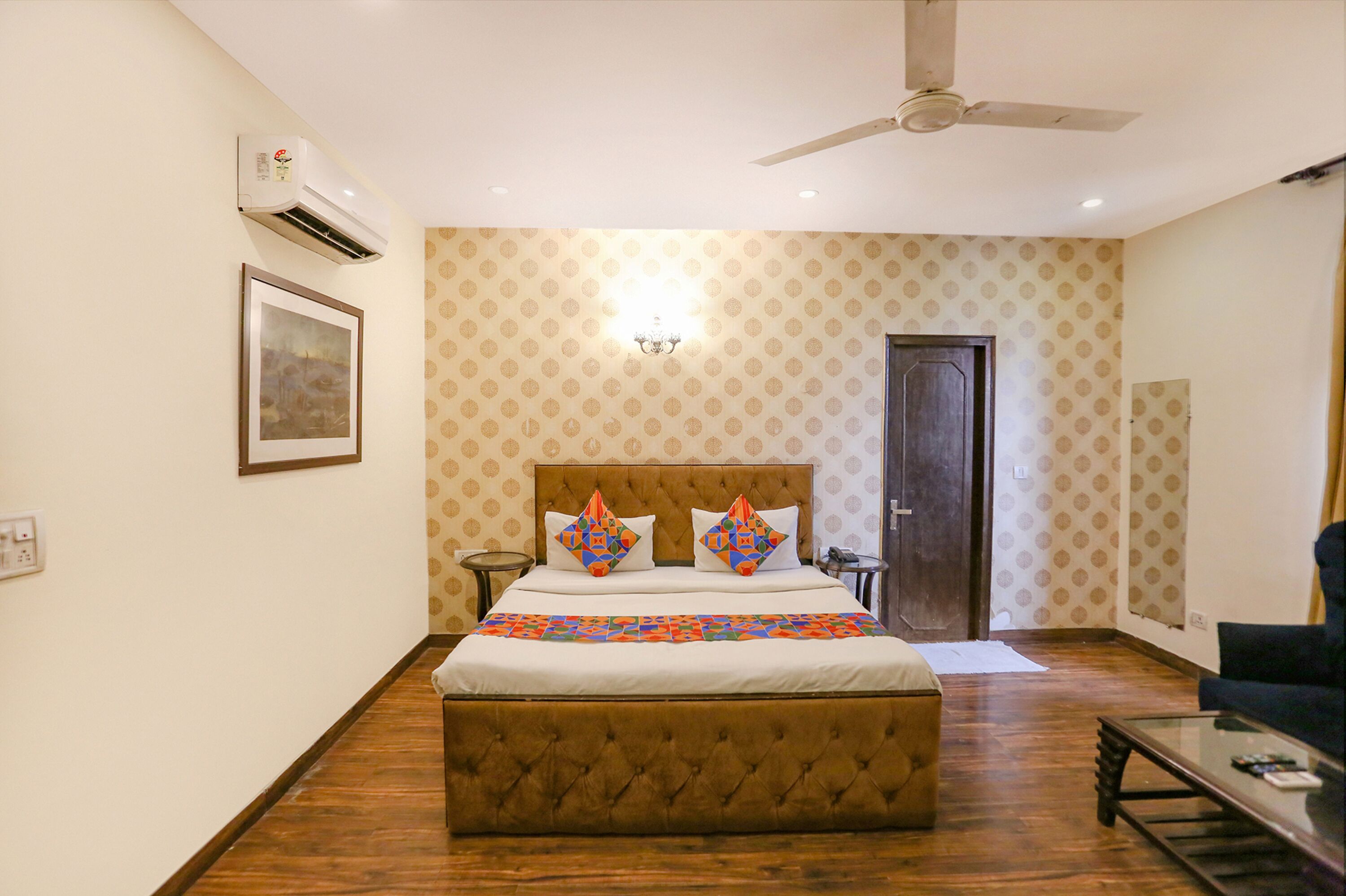 A hotel room with a large bed, a ceiling fan, an air conditioning unit, a framed picture on the wall, and a door.