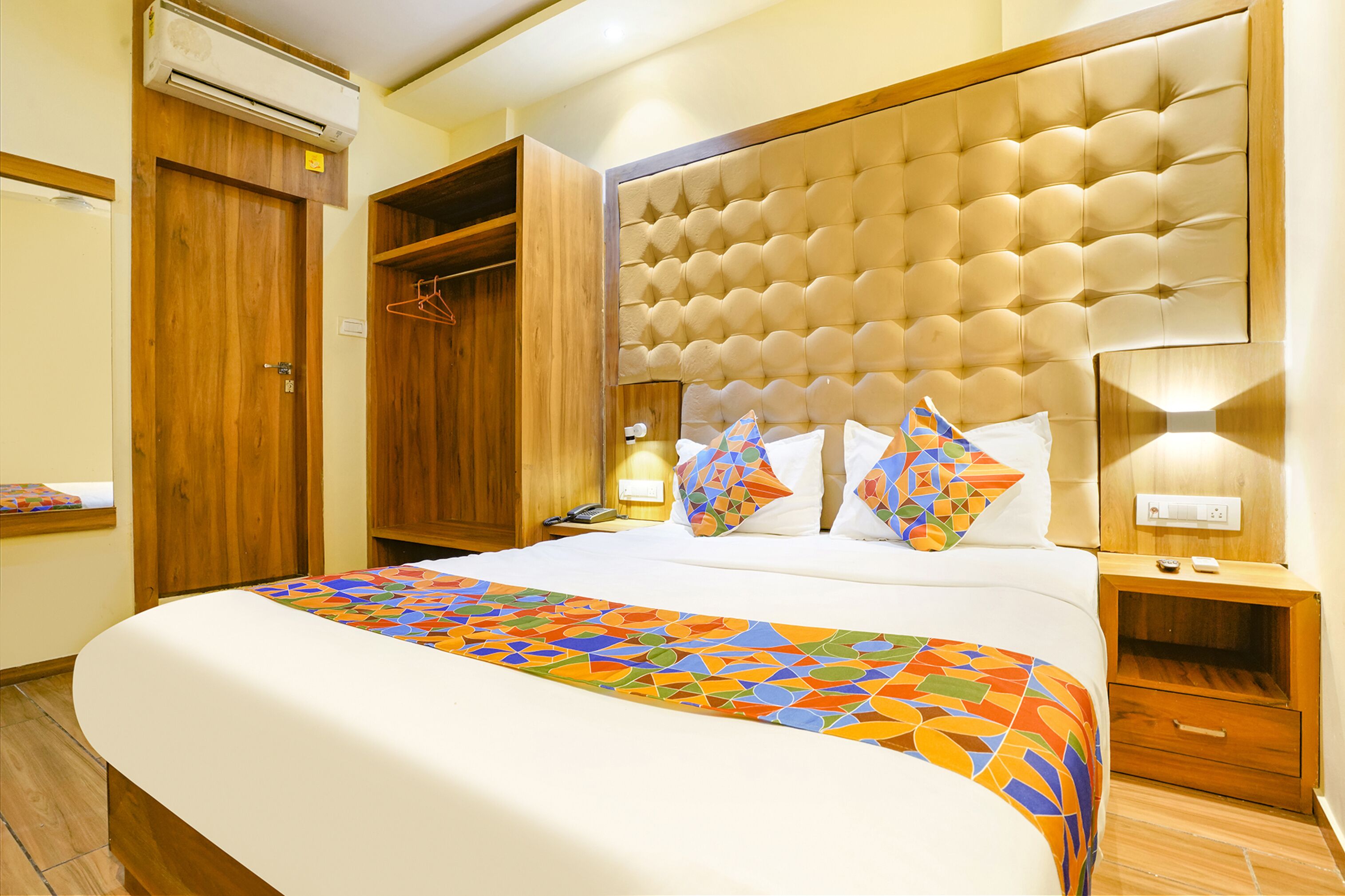 A hotel room with a large bed, a wooden wardrobe, a built-in headboard, and a bedside table with a lamp.