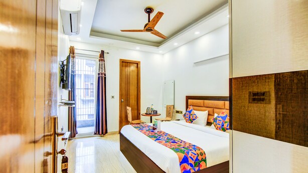 A hotel room with a large bed, a ceiling fan, a wooden door, a small table with a bottle, and a window with curtains. - Fabhotel Floyd (New Delhi)