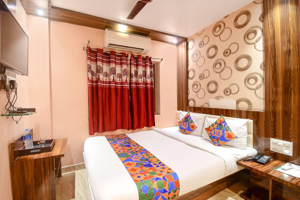 A hotel room with a bed, a bedside table with a phone, a wall-mounted TV, and a red curtain.