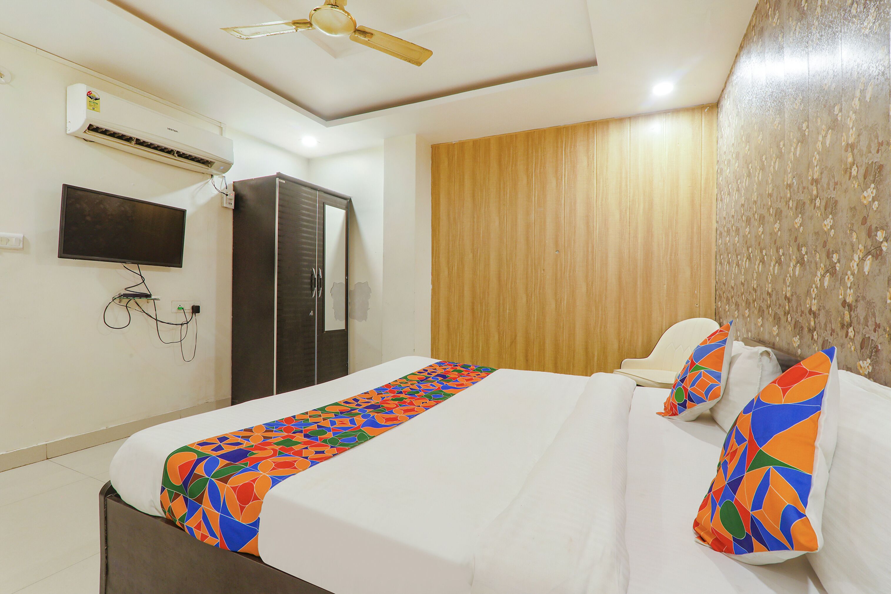 A hotel room with a bed, a television mounted on the wall, an air conditioning unit, a wardrobe, and a ceiling fan.