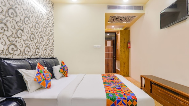 A hotel room with a bed, a black leather sofa, a wooden bench, a patterned wall, and a television mounted on the wall. - Fabhotel Shree Pushpraj (Kanpur)