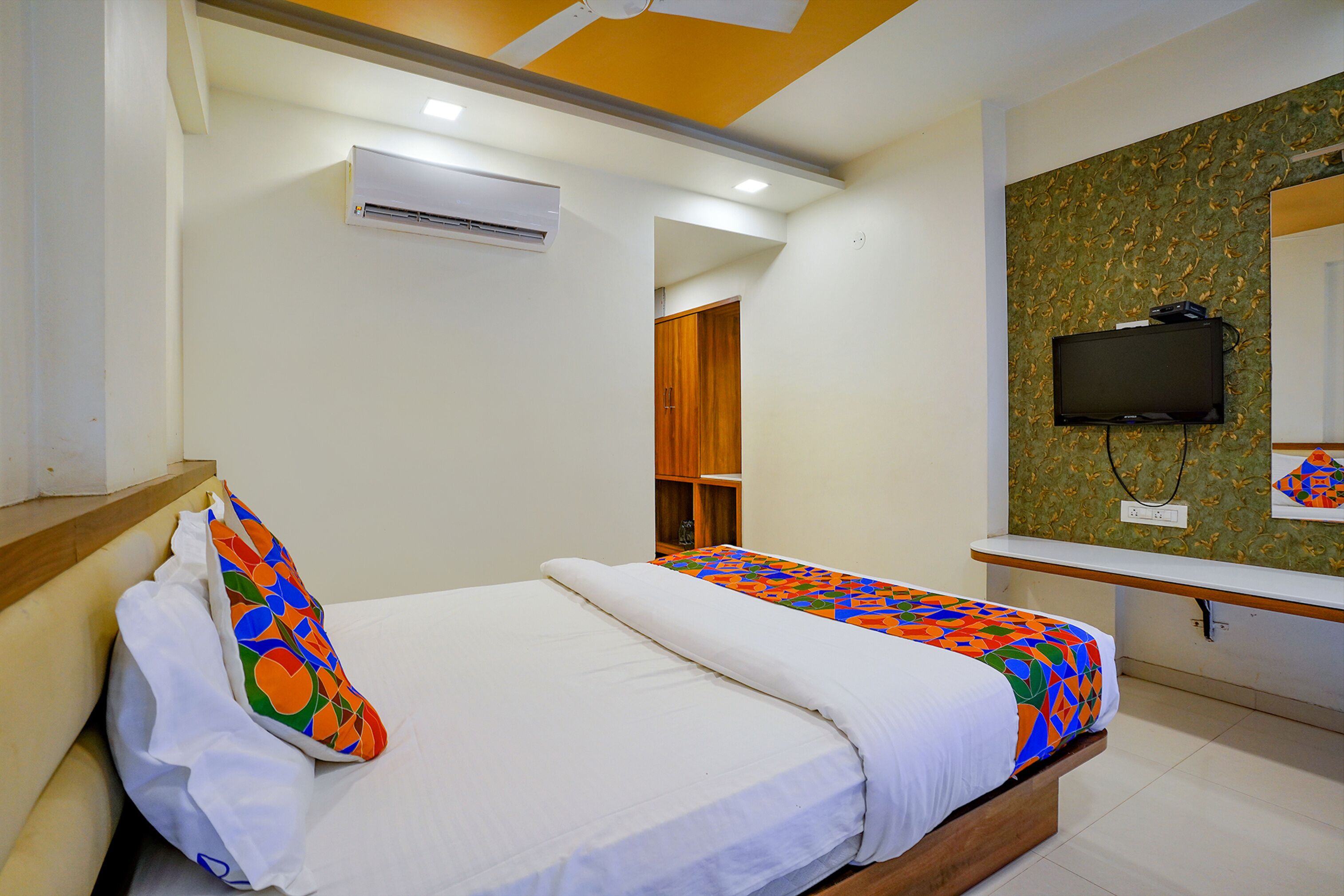 A hotel room with a bed, a TV mounted on the wall, and an air conditioning unit.