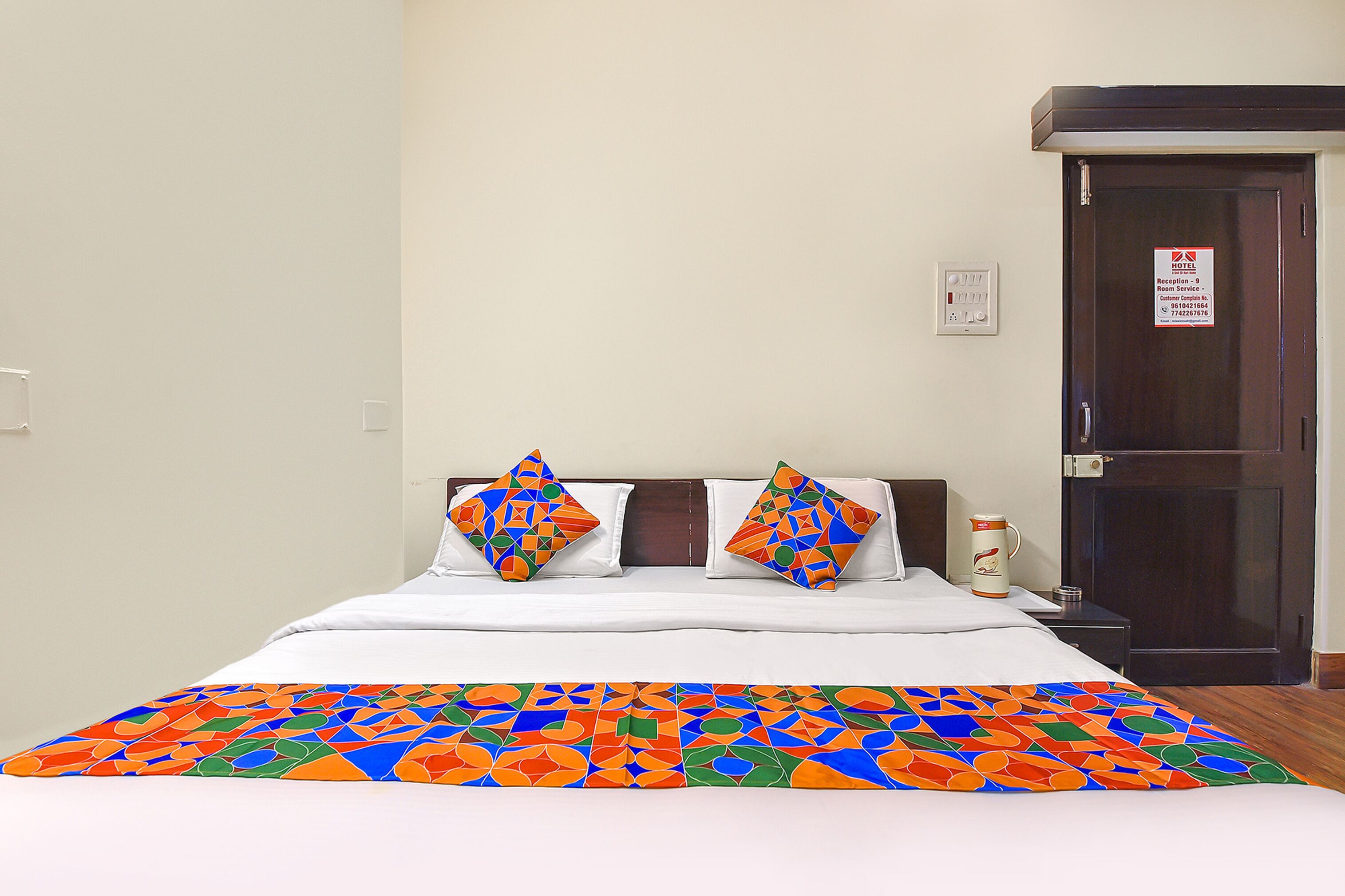 A hotel room with a bed, two pillows, a colorful blanket, a nightstand, a coffee maker, and a door with a sign.