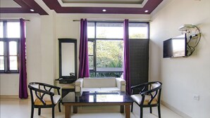 Una habitación con sofá, dos sillones, una mesa, televisor montado en la pared y una ventana con cortinas.