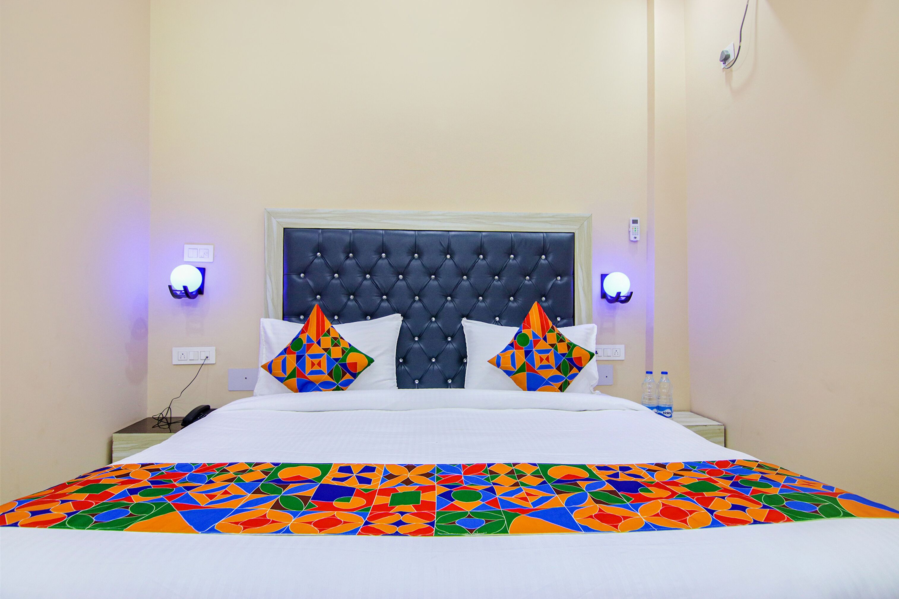 A neatly made bed with a tufted headboard, two decorative pillows, a colorful geometric-patterned bedspread, and two wall-mounted nightstands with lamps.
