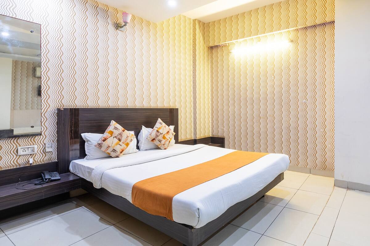 Ein ordentlich bezogenes Bett mit brauner Kopfteil, weißer Bettwäsche und einer orangen Tagesdecke. Ein Nachttisch mit Telefon und Lampe ist sichtbar. Der Raum hat raumhohe Tapete mit Muster und einen großen Spiegel an der Wand.