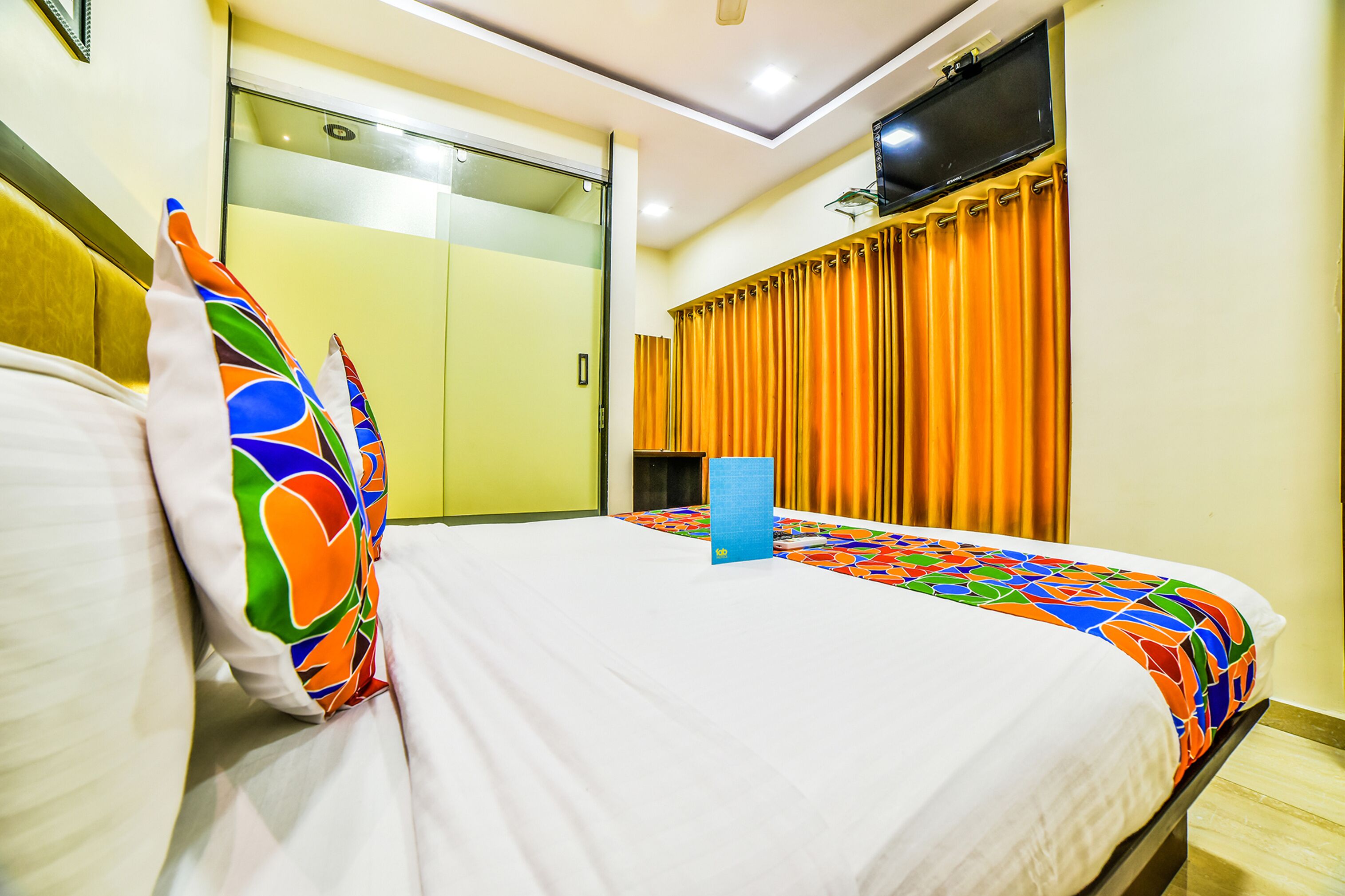 A hotel room with a bed, colorful pillows, a blue bedside table, and a TV mounted on the wall.