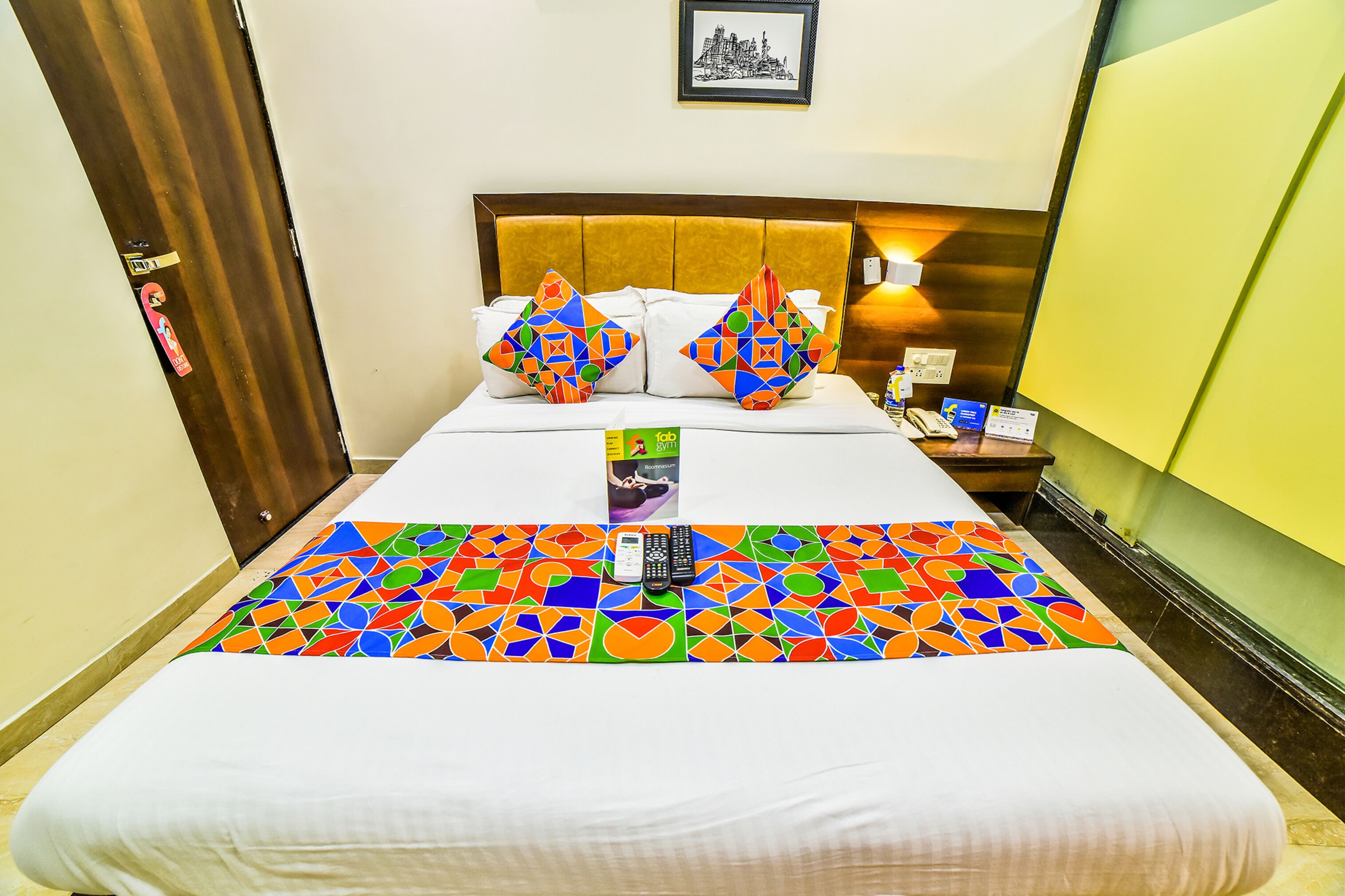 A hotel room with a bed, a nightstand, a framed picture on the wall, and a colorful geometric-patterned bedspread.