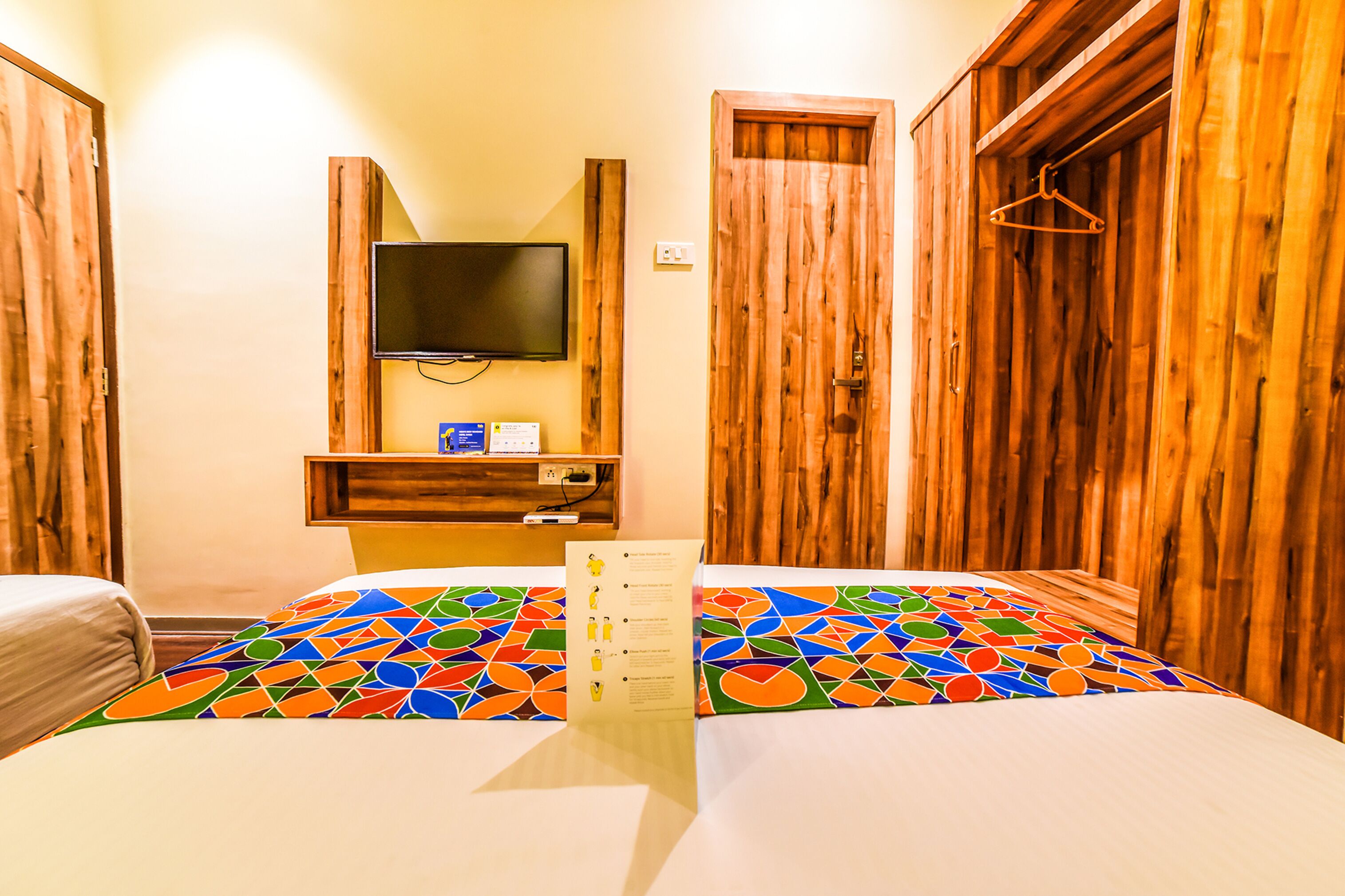 A hotel room with wooden walls, a bed with colorful geometric bedding, a flat-screen TV, and a wooden wardrobe with a hanger.