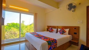 A hotel room with a large bed, a bedside table, a wall-mounted fan, and a window with a view of greenery. - Fabhotel Gr Lake View (Bengaluru)