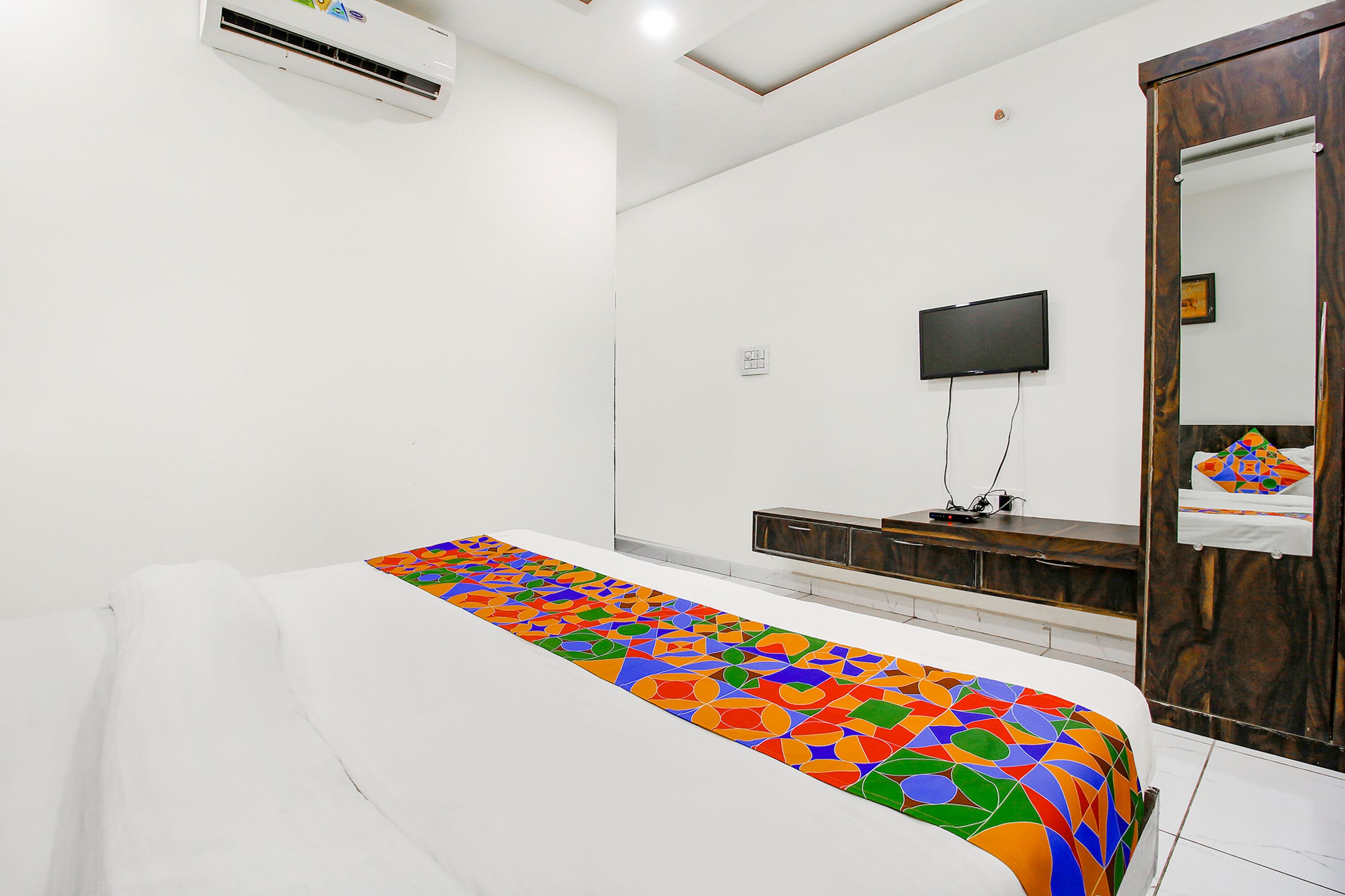 A modern bedroom with a white bed, a colorful geometric-patterned bedspread, a flat-screen TV, and a wooden wardrobe.