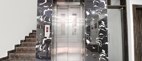 An elevator with a metallic finish, a control panel, and a staircase with dark marble steps.