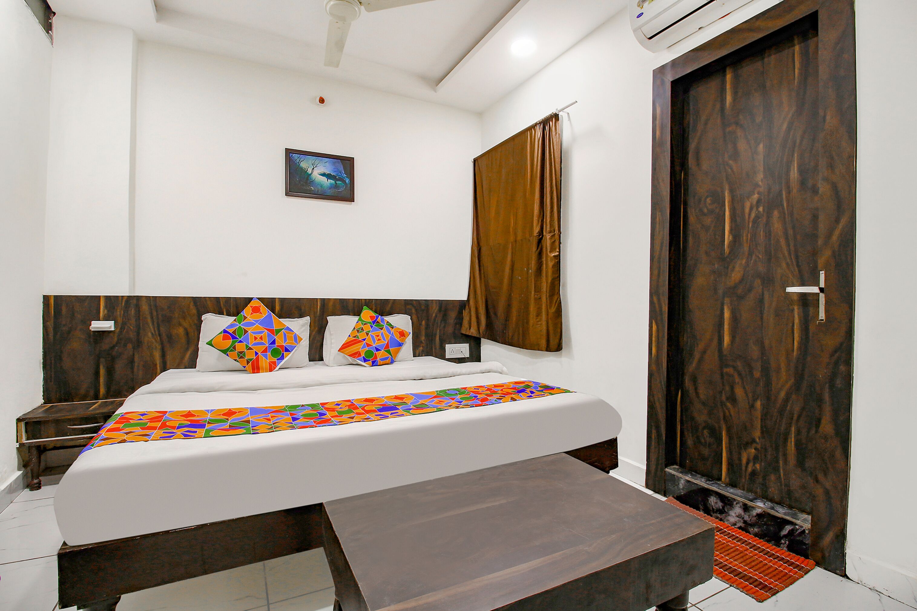 A bedroom with a bed, a nightstand, a wooden headboard, a brown door, and a small picture on the wall.