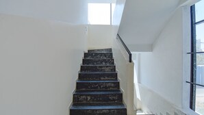 Un escalier avec des marches noires menant à un palier, des murs blancs et une grande fenêtre sur la droite.