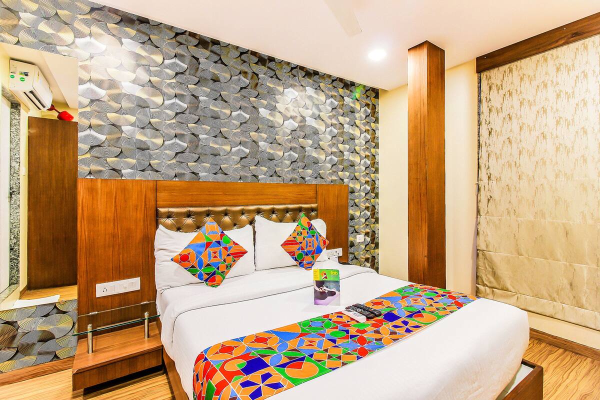 Un lit bien fait, agrémenté d’une couvre-lit aux motifs géométriques colorés, d’une tête de lit en bois et d’un mur décoratif orné d’un motif rappelant des feuilles.
