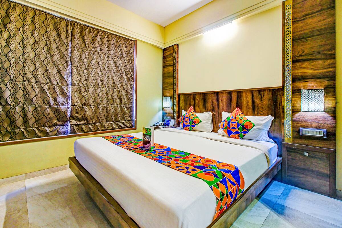 A hotel room with a large bed, a window with a patterned blind, a wooden headboard, and a nightstand with a lamp.