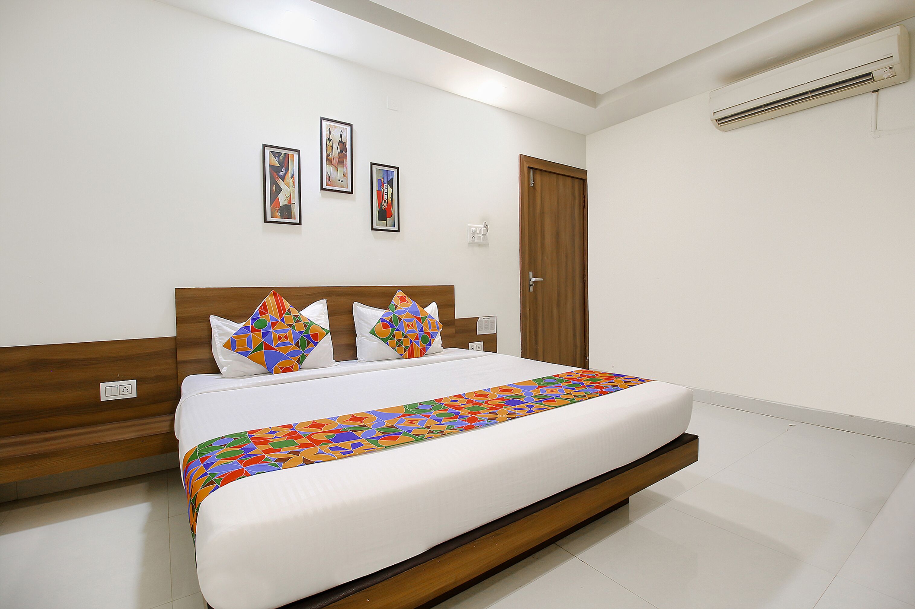 A neatly made bed with a colorful geometric-patterned bedspread, a wooden headboard, and two wall-mounted pictures.