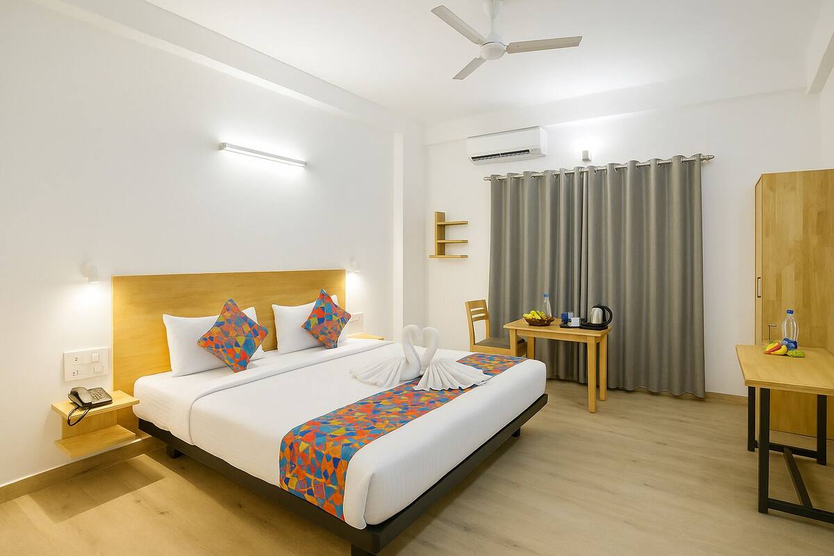 A hotel room with a bed, a bedside table with a phone, a chair, a small table with a kettle, and a mirror.