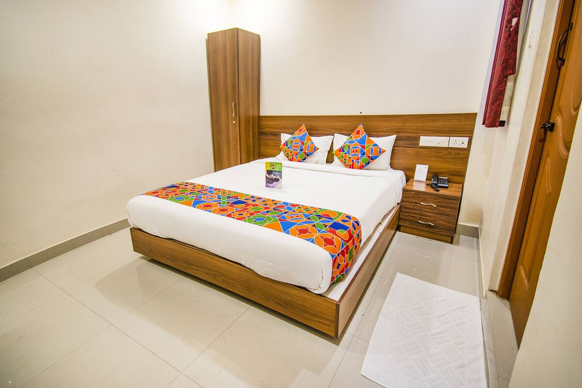 Una cama bien hecha con una colcha de diseño geométrico y colorido y almohadas a juego, un armario de madera, una mesita de noche con una lámpara y una alfombra blanca.