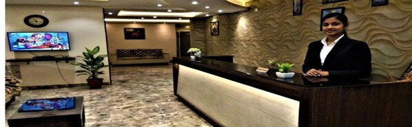 Zona de recepción de hotel con mostrador, televisor montado en la pared y área de descanso con sofá y sillones.
