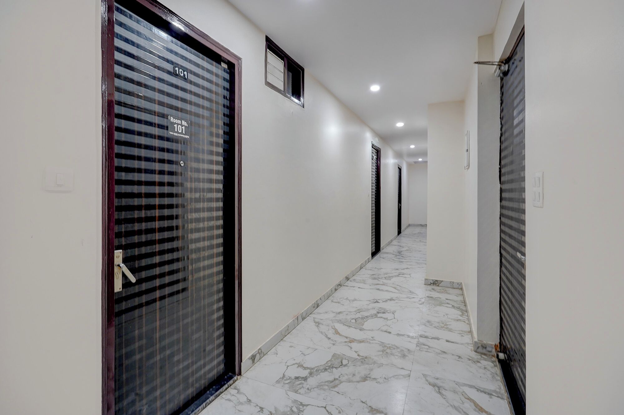 A hallway with a marble floor, doors with room numbers, and recessed ceiling lights.