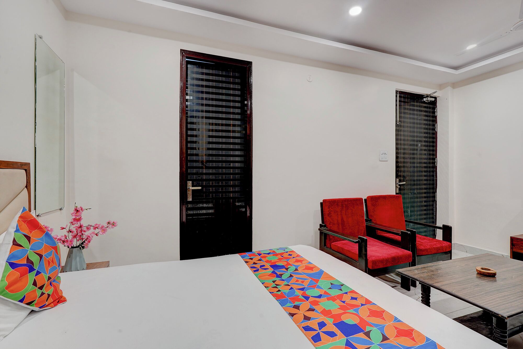 A hotel room with a bed, a table, two red chairs, a vase with flowers, and two black doors.
