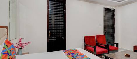 A hotel room with a bed, a table, two red chairs, a vase with flowers, and two black doors.