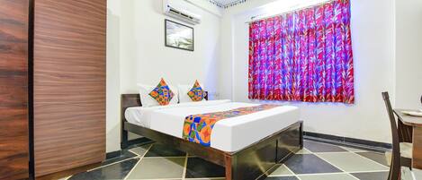 A bedroom with a bed, a wooden wardrobe, a ceiling fan, a window with a colorful curtain, and a tiled floor.