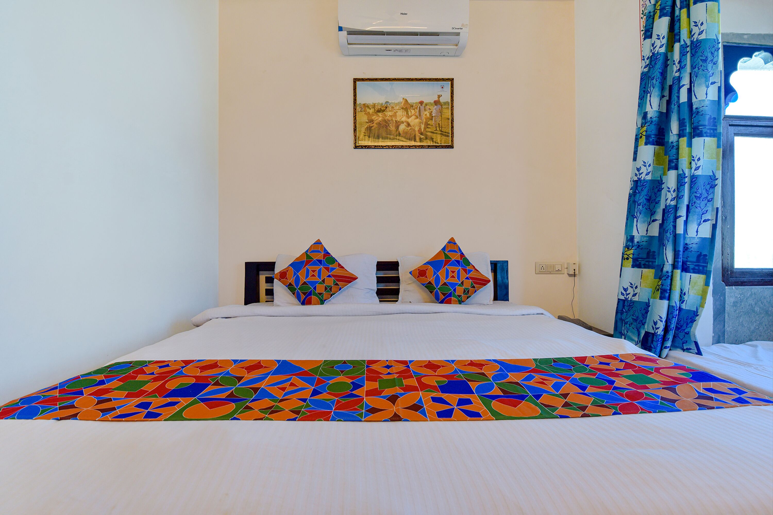 A hotel room with a bed, colorful bedspread, a painting on the wall, and a window with curtains.
