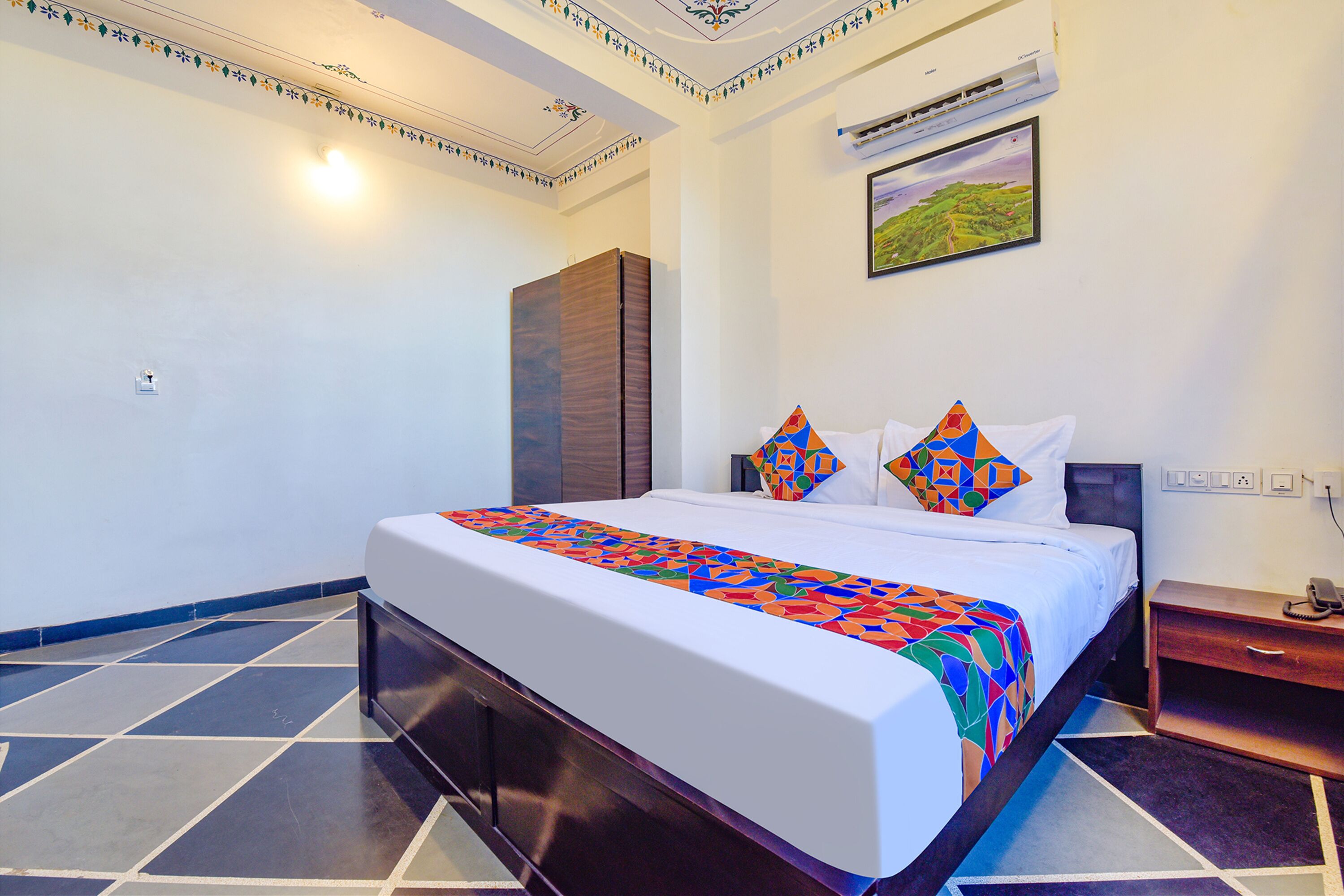 A hotel room with a large bed, a nightstand, a wardrobe, a painting on the wall, and a tiled floor.