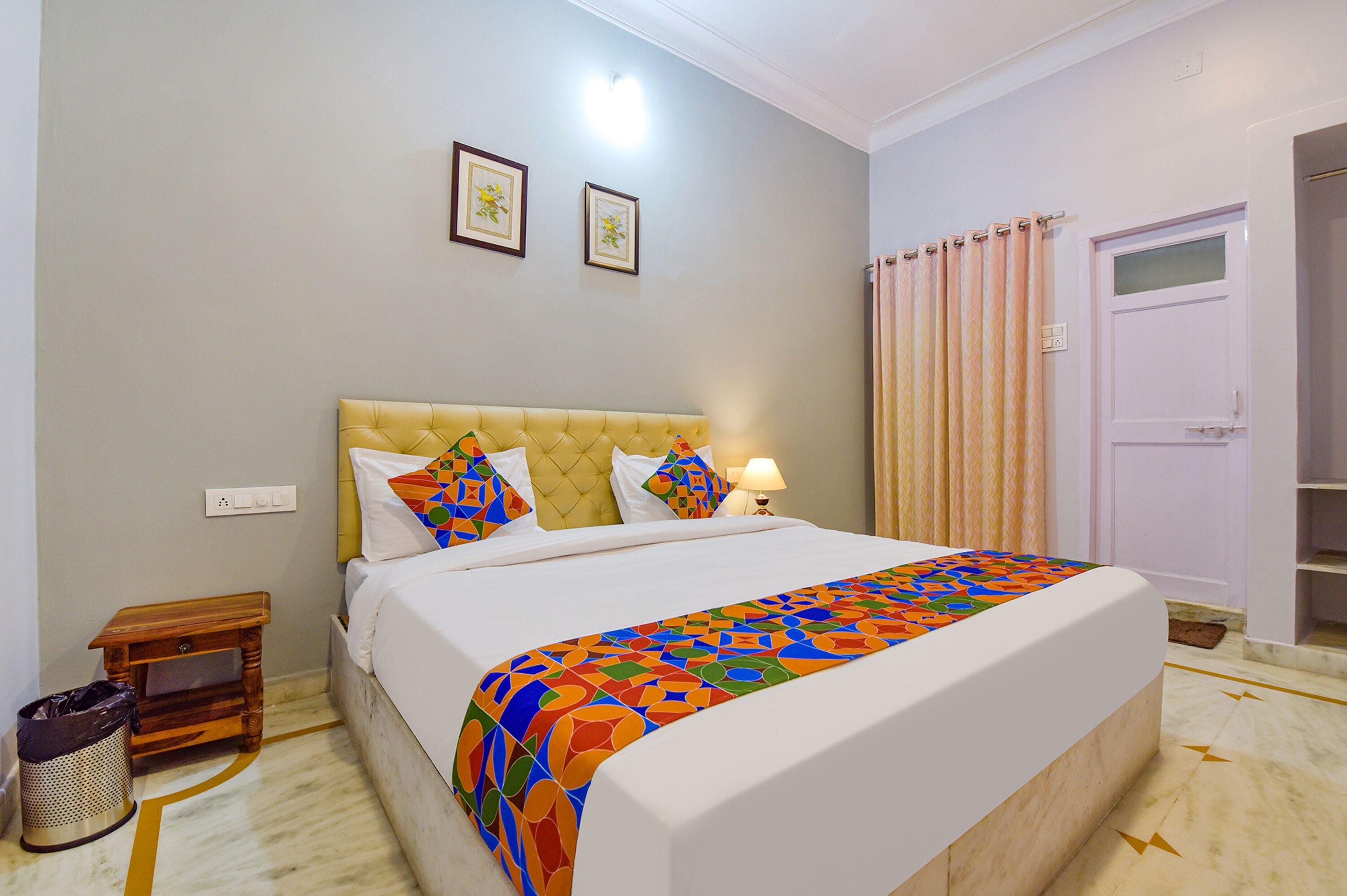 A neatly made bed with a colorful blanket, a bedside table with a lamp, and a trash can.