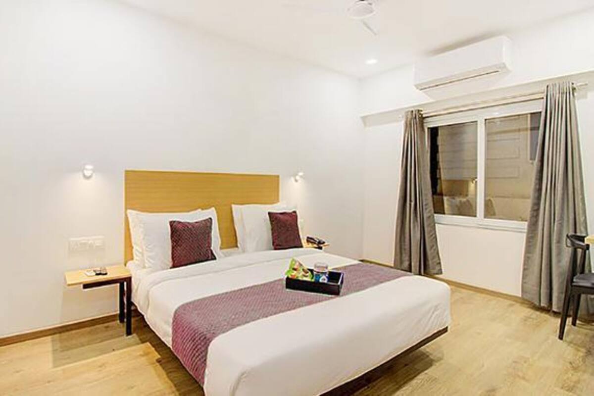 A hotel room with a large bed, a bedside table, a nightstand, a window with curtains, and an air conditioning unit.