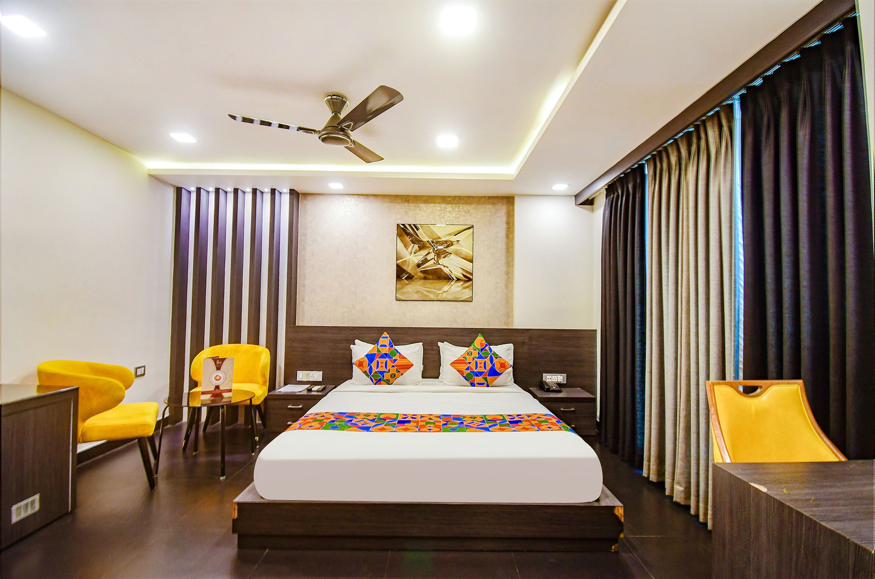 A hotel room with a bed, two chairs, a desk, a ceiling fan, and a large window with curtains.