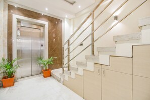 Un ascensor moderno con pared de mármol, una escalera con barandillas metálicas y plantas en macetas.