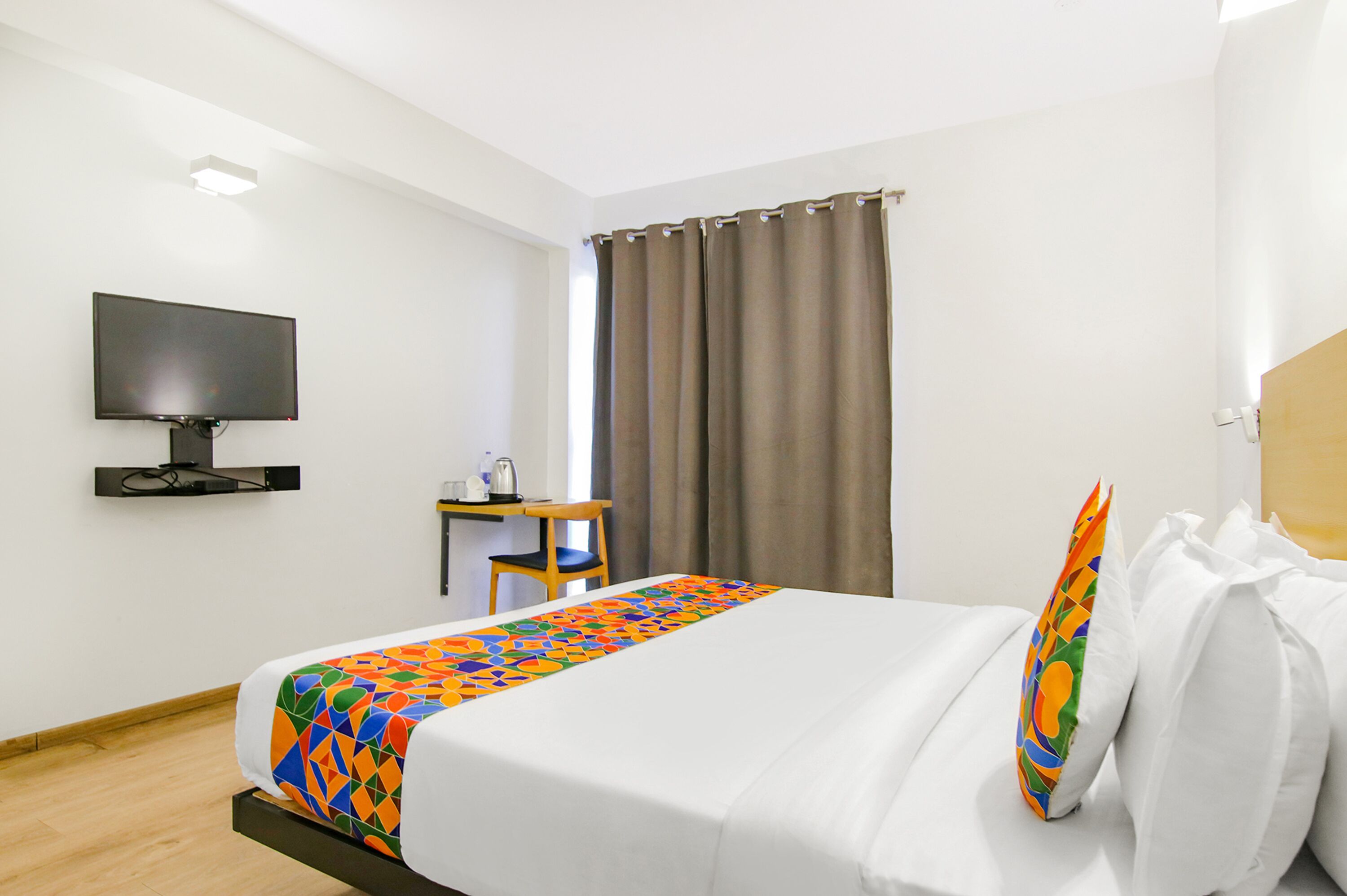 A hotel room with a bed, a TV on the wall, a desk with a chair, and a window with curtains.