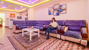Uma sala de estar moderna com um sofá seccional roxo, uma mesa de centro branca e um tapete estampado. Há quadros abstratos na parede e um ventilador de teto.
