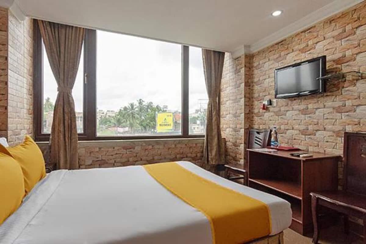 A hotel room with a brick wall, a bed with yellow bedding, a TV mounted on the wall, and a window with curtains.