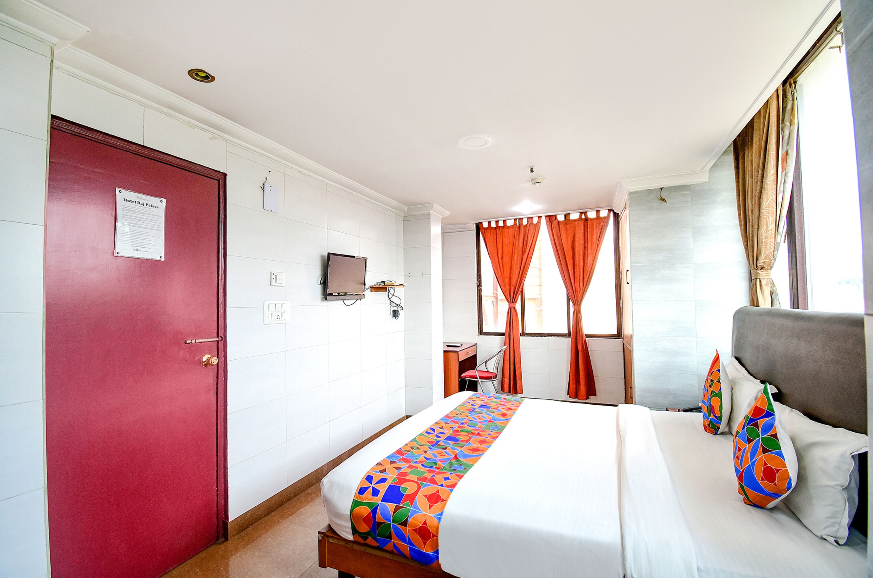 A hotel room with a red door, a bed with a colorful geometric-patterned bedspread, two pillows with a similar design, an orange curtain, and a television mounted on the wall.