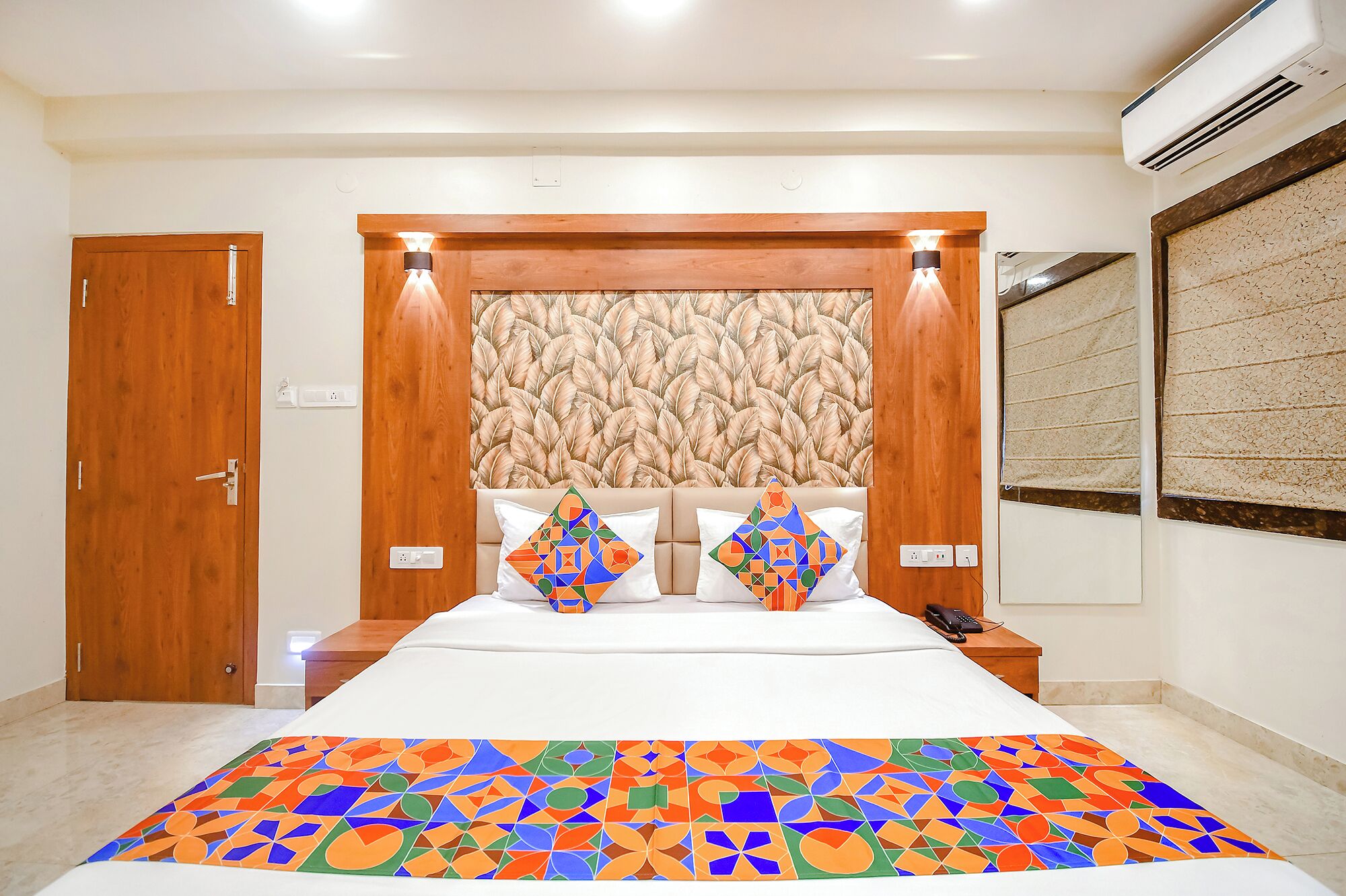 Camera d'albergo con testiera in legno, parete decorata con motivi, un letto con copripiumino geometrico dai colori vivaci e un comodino con un telefono.